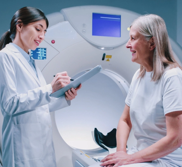 A medical professional writes on a clipboard while facing an elderly woman seated in front of an MRI machine.