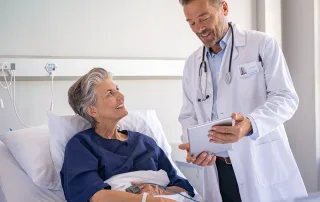 A doctor holding a tablet discusses internal tumor motion with a smiling patient lying in a hospital bed, both appearing engaged in conversation in a clinical setting.